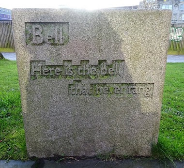 Text: "Bell / Here is the bell / that never rang" carved in shallow relief on a (possibly granite) cuboid stone block.