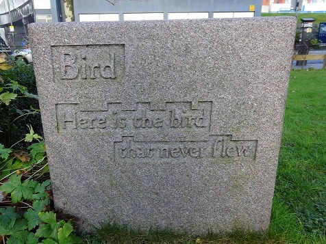 Text: "Bird / Here is the bird / that never flew" carved in shallow relief on a (possibly granite) cuboid stone block.