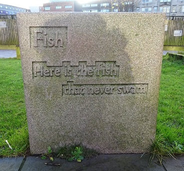 Text: "Fish / Here is the fish / that never swam" carved in shallow relief on a (possibly granite) cuboid stone block.