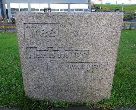 Text: "Tree/ Here is the tree / that never grew" carved in shallow relief on a (possibly granite) cuboid stone block.
