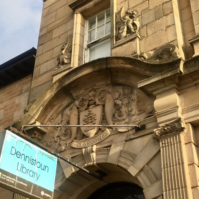 Exterior of the entrance to Dennistoun Library/ Victorian yellow sandstone with arch above doorway which in turn is topped by a formal Glasgow Coat of Arms in relief. A modern sign sticks out from the inside top of the arch with text showing the library's name