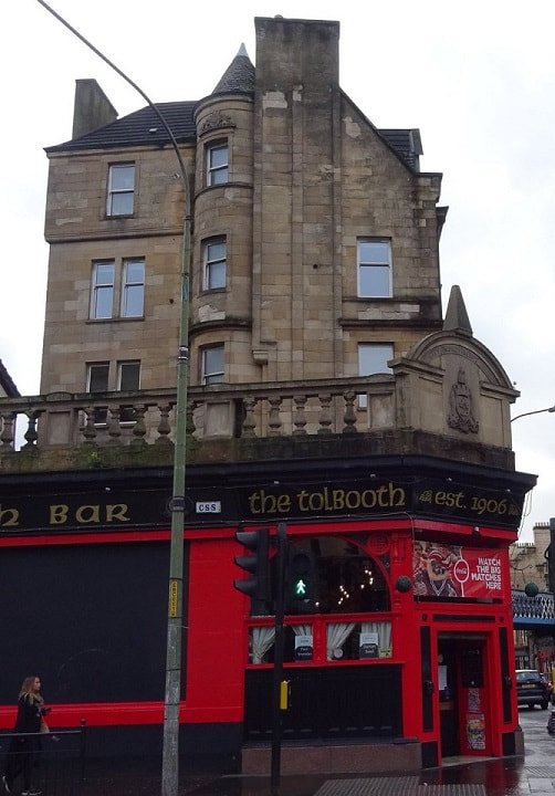 Photo of the Tolbooth Bar at Glasgow Cross (11 Saltmarket). There's a City Improvement Trust Glasgow CoA atop the corner entrance. A sandstone building gable end looms behind the pub