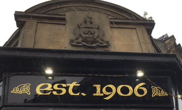 Formal Glasgow coat of arms relief above the corner doorway of the Tolbooth Bar, 11 Saltmarket. Image shows text below the CoA saying "est. 1906". There's an arched band above the CoA with text, "City Improvement Trust"